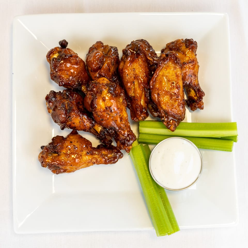 A plate of glazed chicken wings with celery sticks and a side of ranch dressing.