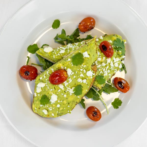 Two avocado toasts with feta, roasted tomatoes, and cilantro on a white plate.