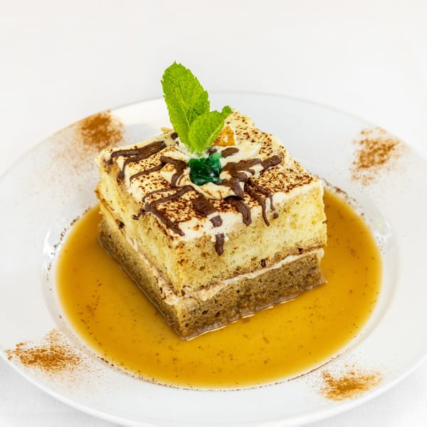 A slice of tiramisu cake with chocolate drizzle, mint, and caramel sauce on a white plate.