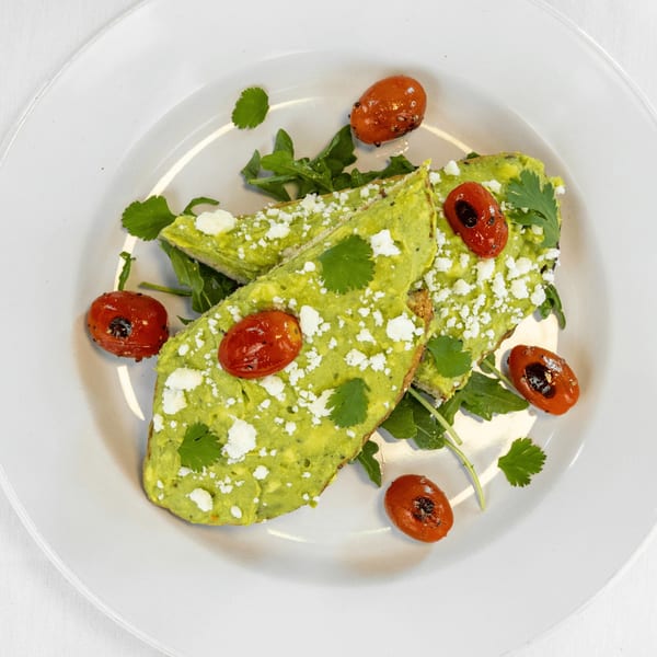 Avocado toast with feta, roasted tomatoes, and cilantro on a white plate.