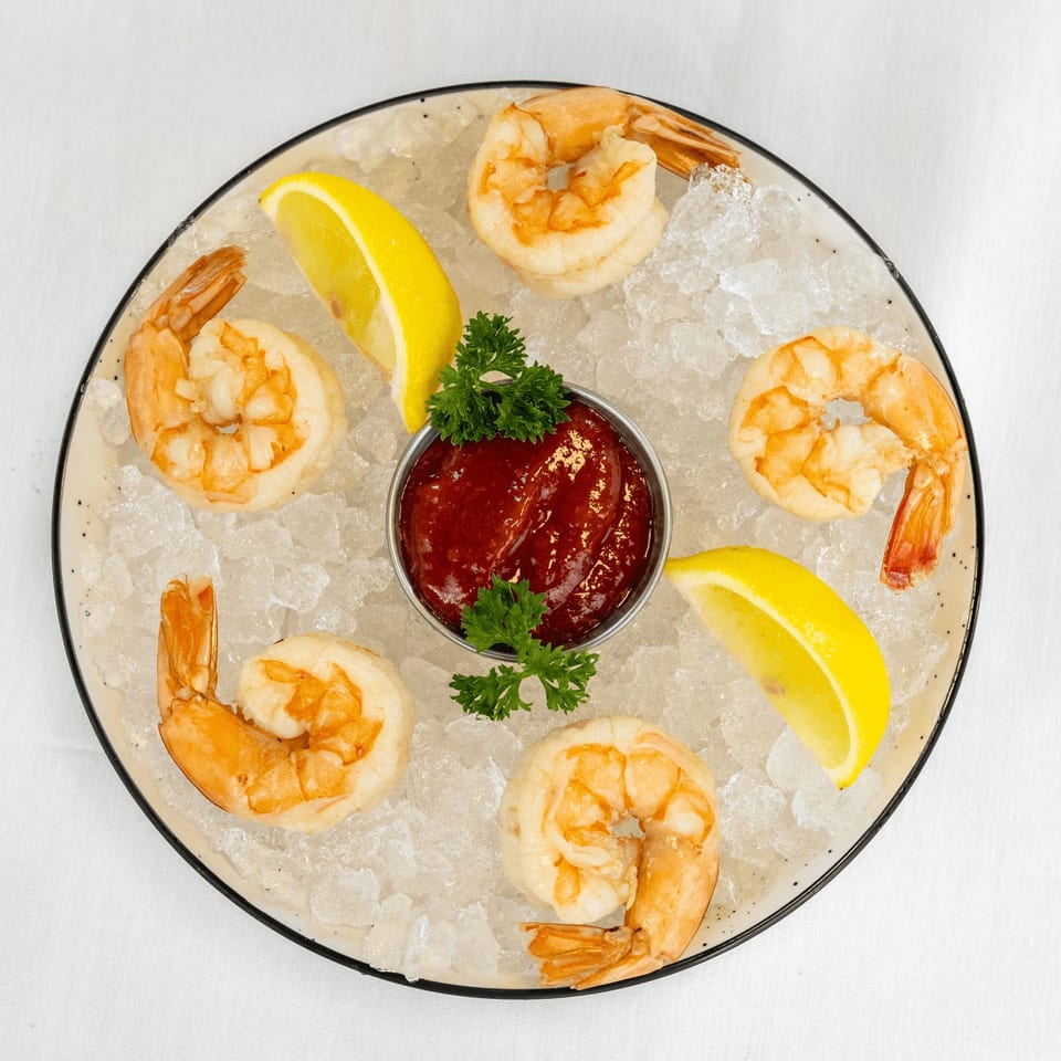 Shrimp cocktail with six large shrimp, cocktail sauce, lemon wedges, and parsley on a bed of ice.