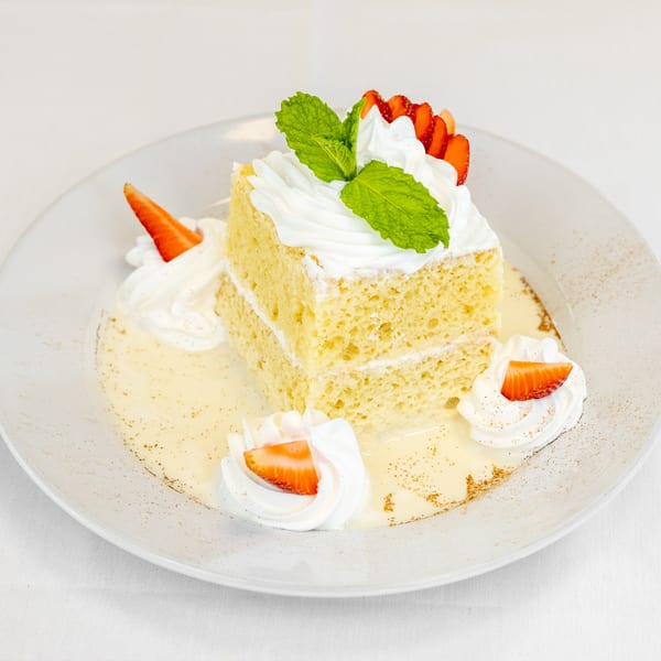 A slice of Tres Leches cake with whipped cream, strawberries, and mint on a white plate.