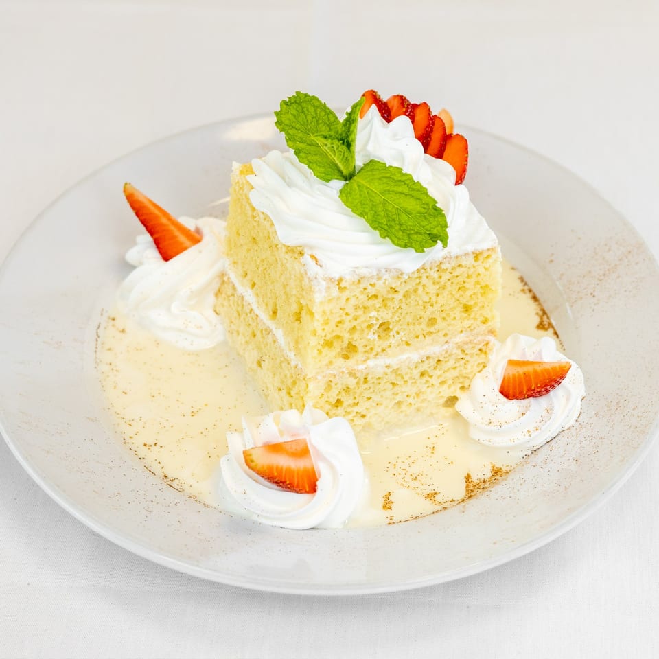 A slice of Tres Leches cake with whipped cream, strawberries, and mint on a white plate.