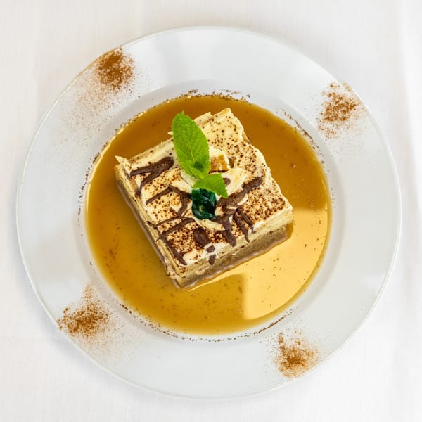 A square dessert with cream, chocolate drizzle, and mint, in a caramel sauce on a white plate.