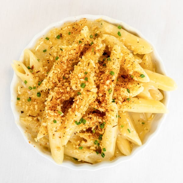 Creamy penne pasta with toasted breadcrumbs and chives in a white bowl on a white tablecloth.